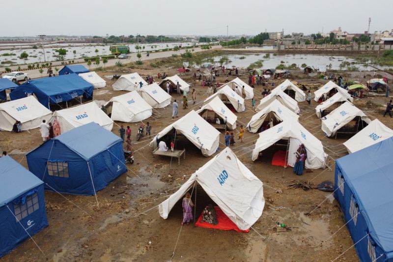 Blaue und weiße Zelte einer Hilfsorganisation sind in Reihe aufgebaut, im Hintergrund sind Felder weithin überflutet.