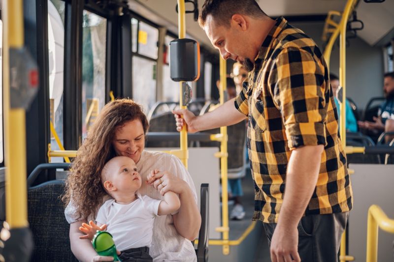 Ein Mann im Holzfällerhemd steht in einem Bus und spricht mit einem Kleinkind, das auf dem Schoß der Mutter sitzt.