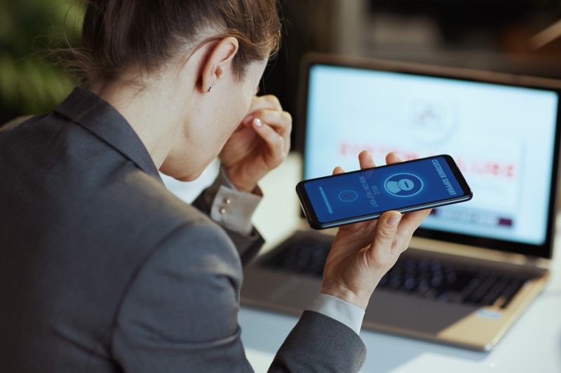 Eine gestresste Geschäftsfrau mit Laptop und Smartphone will den Kundendienst kontaktieren und erreicht nur einen Chatbot.