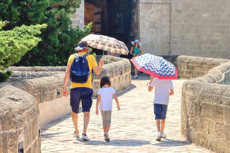 Ein Mann und zwei Kinder gehen einen Steinweg entlang, zum Schutz vor der Sonne haben sie Regenschirme aufgespannt. Eine Aufnahme aus Apulien in Süditalien.