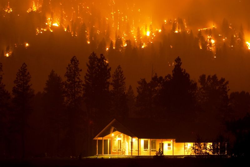 Ein einstöckiges, nächtlich beleuchtetes Holzhaus am Waldrand, in den Bergen dahinter brennt der Wald, es sieht aus der Ferne wie Feuerwerk aus.