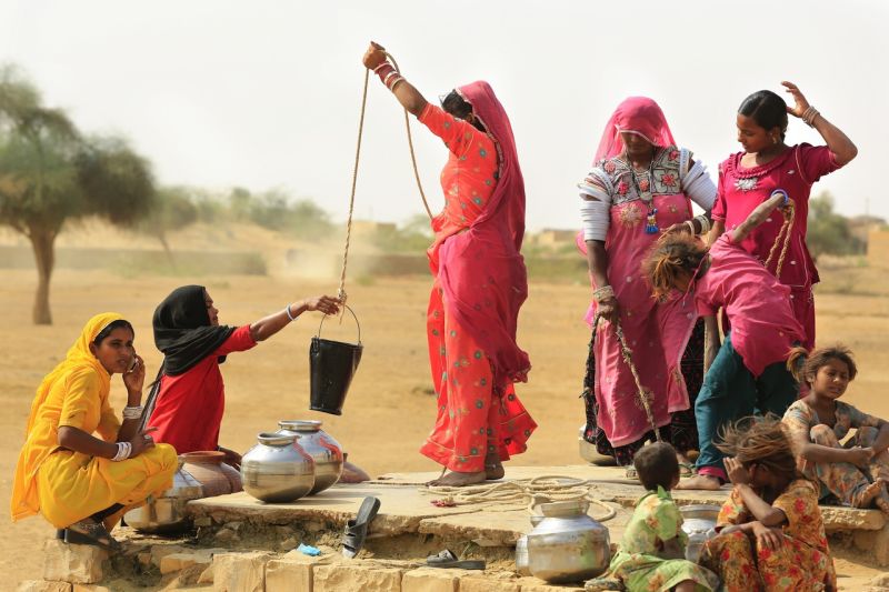 Frauen in roten und gelben Saris und einige Kinder an einer Wasserstelle, der gefüllte Eimer wird gerade hochgezogen, Blechgefäße stehen bereit.