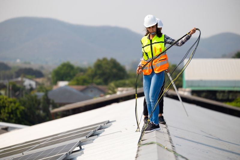Eine Frau und ein Mann in Arbeitskleidung balancieren auf einem Dach, auf dem Solarpaneele installiert werden. 