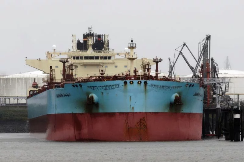 Ein in die Jahre gekommenes Tankschiff in verblichenem Blau und Rot mit weißen Aufbauten ist fast genau von vorn zu sehen, daneben Hafenanlagen.
