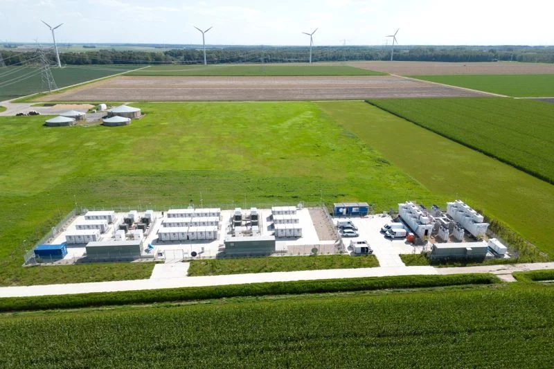 Weiße Container mit Batteriespeichern stehen auf einem Feld, im Hintergrund ein Windpark und einige Biogasspeicher.