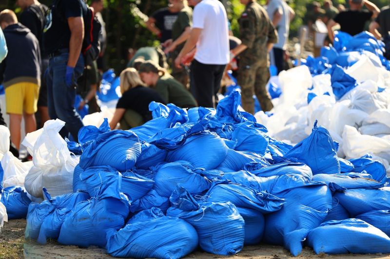 Blaue und weiße Plastiktüten-Sandsäcke liegen auf einem Haufen, dahinter sind Menschen mit dem Füllen beschäftigt.