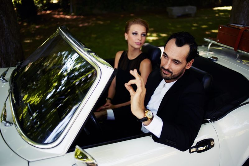 Ein in festliches Schwarz gekleidetes Paar im jüngeren bis mittleren Alter sitzt in einem Luxus-Cabrio. Der Mann, hinter dem Lenkrad, macht eine Okay-Handbewegung, er trägt eine teure Uhr.