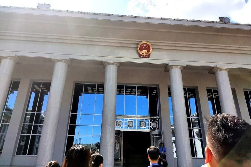 Einige Menschen stehen vor einem weißen Monumentalbau mit Säulen, großen Fensterfronten und dem Zeichen der Obersten Staatsanwaltschaft.