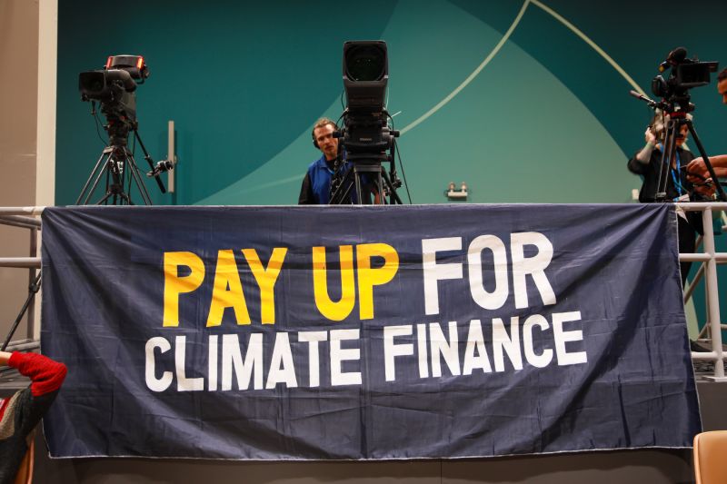 Protestbild und Kameras auf dem Klimagipfel 2024 in Baku. Auf einem Transparent der Slogan auf Englisch: Bezahlt für die Klimafinanzierung!