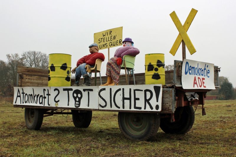 Bauer und Bäuerin sitzen als lebensgroße Strohpuppen auf einem Lastwagen-Anhänger, neben ihnen zwei als Atommüll angemalte Fässer, ein gelbes X aus Holz und mehrere Schilder: Atomkraft? Todsicher! – Demokratie ade. – Wir stellen uns quer!